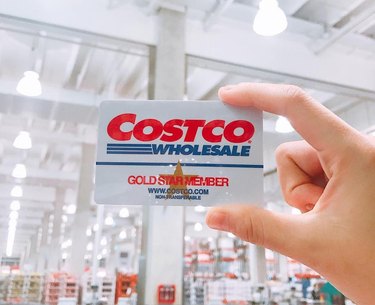 A light-skinned hand hold up a Costco gift card in front of a well-lit Costco warehouse.
