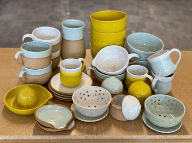 Stacks of mugs, bowls, and colanders in light blue, white, gray, and yellow on a wooden surface.