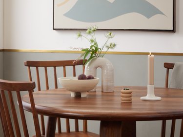 A dining room wood table topped with an arched ceramic white vase, a light brown wavy match holder, a sculptural fruit food, and a white candlestick holder with a tan tapered candle in it.