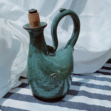A greenish blue ceramic drink canister on a striped blanket in front of a white sheet.