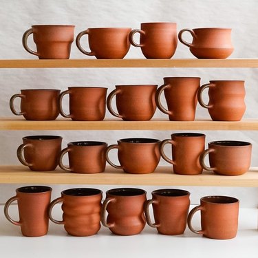 Four wooden shelves with brick-colored mugs all in different shapes in front of a white wall.