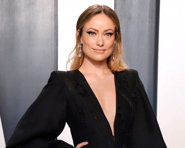 Olivia Wilde waist up posing with her hand on her hip wearing a long sleeve black dress with a v-cut neck.