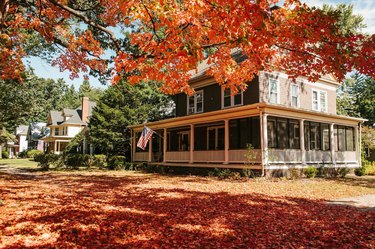 Exterior of a beige house with a wraparound porch behind color fall leaves.