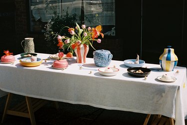 An outdoor dining table featuring a red striped vase of flowers, plates with primary color rims, and speckled jars.