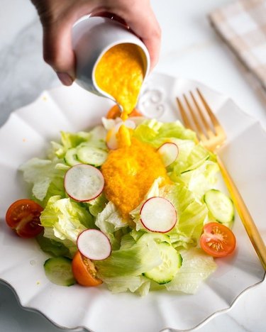 A simple salad of lettuce, cucumber, cherry tomatoes, and radish with an orange carrot ginger dressing being poured on top of it.