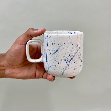 person holding white mug with blue paint splatters