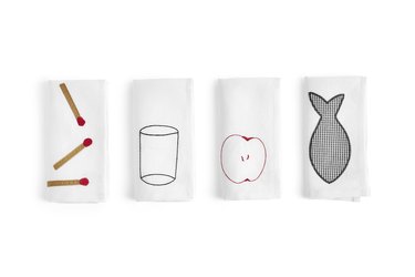 Four white napkins featuring, respectively, embroidered matches, a glass, an apple, and a fish.