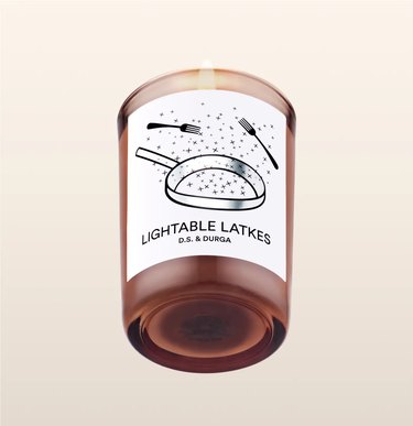D.S. & Durga's Lightable Latkes candle in a transparent brown jar with an illustration of a frying pan and two forks on it.
