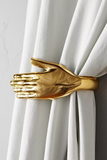 A gold hand curtain holdback holding a white curtain