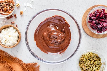 Melted dark chocolate in a glass bowl surrounded by the other ingredients in glass bowls on a white counter.