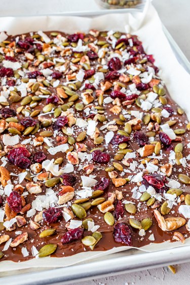 The melted chocolate on the baking sheet topped with dried cranberries, pepitas, coconut flakes, pecans, and sea salt.