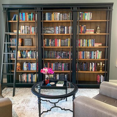 A black bookshelf with a black ladder leaned up against it. There's a black coffee table and a partial view of a beige chair.