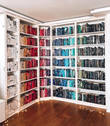 A white floor-to-ceiling bookshelf with books organized by color in order of the rainbow.