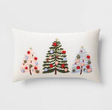 A small white lumbar pillow with three holiday trees on it, one is pink with red, blue and green ornaments, the largest is green and in the middle with red, blue and pink ornaments, and the last one is light blue with green, pink and red ornaments.