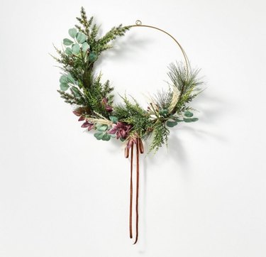 An asymmetrical wreath with faux pine needles mixed with eucalyptus leaves,  mixed with red leaves and a red ribbon tied at the bottom.