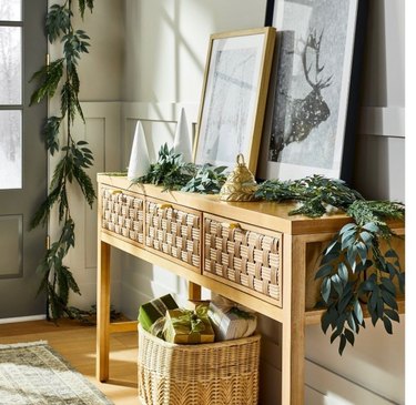 An unlit faux cedar and eucalyptus garland lays on a light brown console table. A basket filled with olive green presents rests underneath the table, with two white trees, a gold bell and two framed winter prints rest on top.