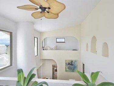 A balcony that overlooks the interior of a white home with arched ceilings and windows, with a brown light fixture on the ceiling that resembles leaves.