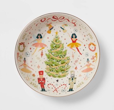 A white serving bowl with a Nutcracker theme. A green Christmas tree is at the center, surrounded by two nutcrackers, four ballerinas, and an array of red ribbons, candy canes, wreaths, trumpets and bells.