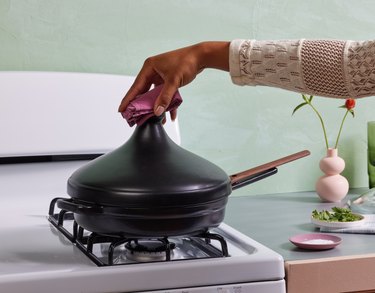 A person holding an black tagine cover over a black Always Pan on a white stove.