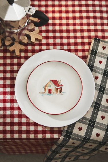 A white plate with a red rim and a gingerbread house illustration in the center of a tablecloth that features red and white checks.