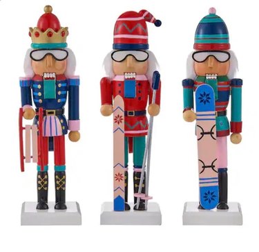 Three nutcrackers, all wearing ski goggles. The first one has a red and gold crown, white hair, a navy blue top with red bottoms and black boots. He's holding a sled. The second is wearing a red hat, has a white hair and is holding ski poles and pink skis with a red snowflake print and blue lines. He wears a red top with green bottoms and black boots. The third nutcracker wears a green and blue hat, has white hair and is holding a pink snowboard with blue ends, featuring a darker blue snowflake. He wears a green, blue and red top, pink bottoms and black boots.