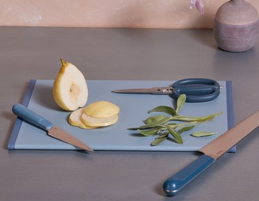 A blue cutting board with a pear and sage on top of it, along with two blue knives and kitchen scissors.