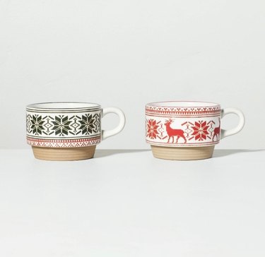 Two small mugs, both with tan bottoms and white handles, the one on the left is green snowflake Fair Isle print, and the one on the right is red reindeer and snowflake Fair Isle print