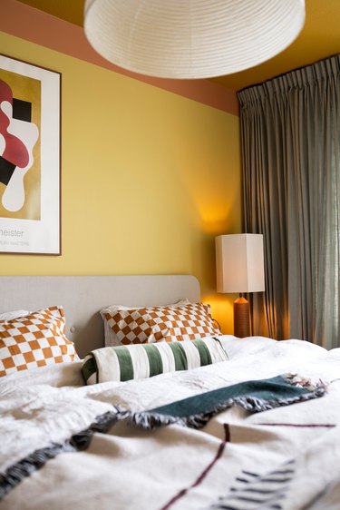 A modern and layered bedroom with orange checkerboard pillows, striped bolster pillow, yellow and orange walls and sage green curtains.