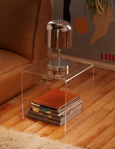 Acrylic end table with metal lamp, books, wood floor.