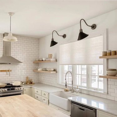 A white kitchen with a large window covered in a cellular shade; black sconces, open shelving, and a farmhouse sink