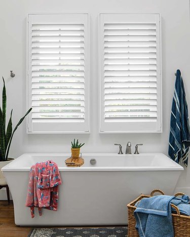 A white bathroom with a freestanding tub and louvered window blinds