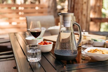 A Hydros water pitcher in gray rests on a black outdoor dining table, surrounded by a glass of red wine, a small glass of water, a white bowl filled with crackers, another white bowl filled with cherry tomatoes, and what looks like a cheeseboard is in the background.