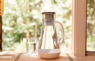 A clear Hydros water pitcher with a white bottom rests on a window sill on a sunny day. There are trees in the background, and a clear glass of water next to the pitcher.