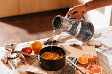 A clear, grey Hydros water pitcher is being held by a medium-toned hand, and water is being poured into a simmer pot, filled with orange slices and sprigs of rosemary. The pot lies on a wooden cutting board surrounded by additional orange slices, apples, and a glass of water with two cinnamon sticks in it. The kitchen counter wis covered with a white and black lined table cloth.