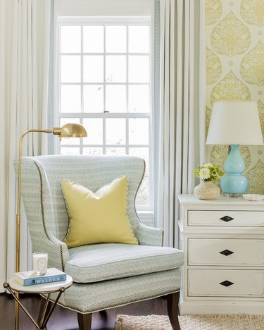 Room corner with pastel blue and white patterned side chair with yellow accent pillow, pale yellow patterned wallpaper and white curtains.