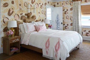 Bedroom with patterned pale yellow, pink and brown wallpaper, beige and white curtains, brown upholstered bed frame, and white and pink bedding .