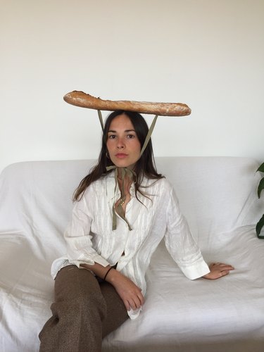 A photo of Sarah Espeute, a person with long brown brain and a baguette tied to their head with green ribbon.