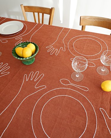 A burnt orange tablecloth on a table embroidered with plates, spoons, and hands in white.