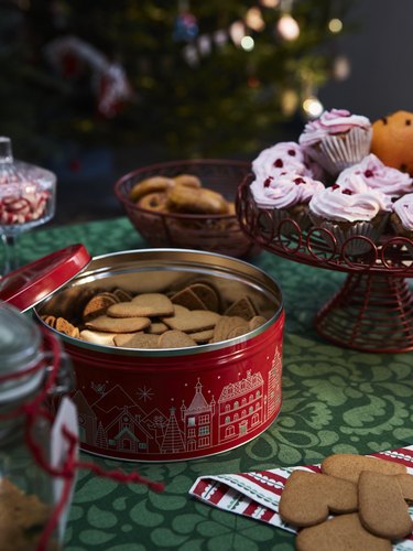 A red tin full of cookies on a green printed table with other sweets in the back like pink-frosted cupcakes and bowls of cookies.