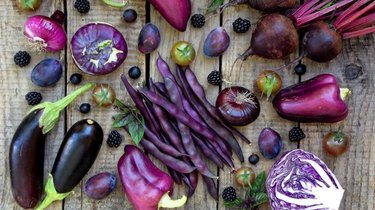 Purple fruits and vegetables, like peppers, cabbage, eggplant, and tomatoes, laying out on a wooden surface