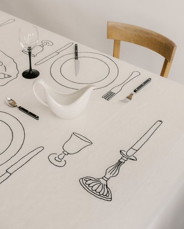 A white tablecloth embroidered with a candlestick, glasses, plates, knives, and forks in black thread.