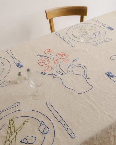 A beige tablecloth featuring embroidered blue plates, forks, knives, snails, spoons, and a vase with pink flowers.