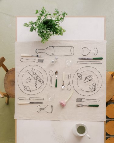 A white table topped with a beige tablecloth with embroidered black plates, forks, knives, glasses, a wine bottle, snails, oysters, a lemon, and asparagus.