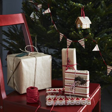 Four wrapped presents on a red chair next to red ribbon in front of a decorated Christmas tree.