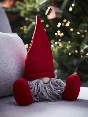 A stuffed gnome with a red hat, red boots, and gray beard on a light gray cushion in front of a Christmas tree.