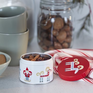 A white tin with drawings of Santa and reindeer full of almonds on a white kitchen counter with mugs in the background.