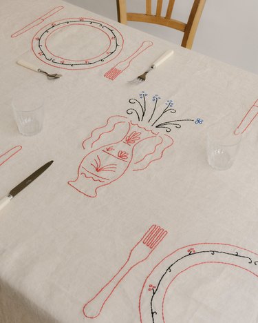 A white tablecloth embroidered with a pink vase of blue flowers, along with plates, forks, and knives in pink and green.