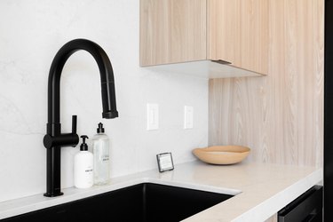 Curved black faucet over a drop-in sink, white countertop, wood kitchen cabinets, and a beige bowl