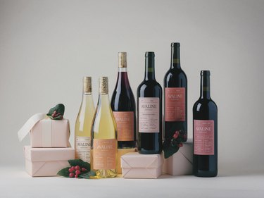 The six wines that make up Avaline's holiday collection rest on an assortment of light pink wrapped gifts. There are holly and berries as well, and the wines include a Viognier, Sauvignon Blanc, Syrah, Bobal, Penedés Tinto, and a Merlot