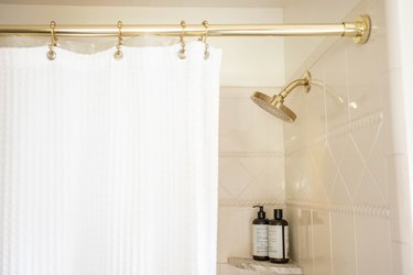 Shower with a gold rod, white shower curtain, gold shower head, and a small corner shelf with two bottles